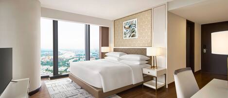 A modern hotel room with a large bed, a desk, a chair, a TV, and a view of the city through large windows.