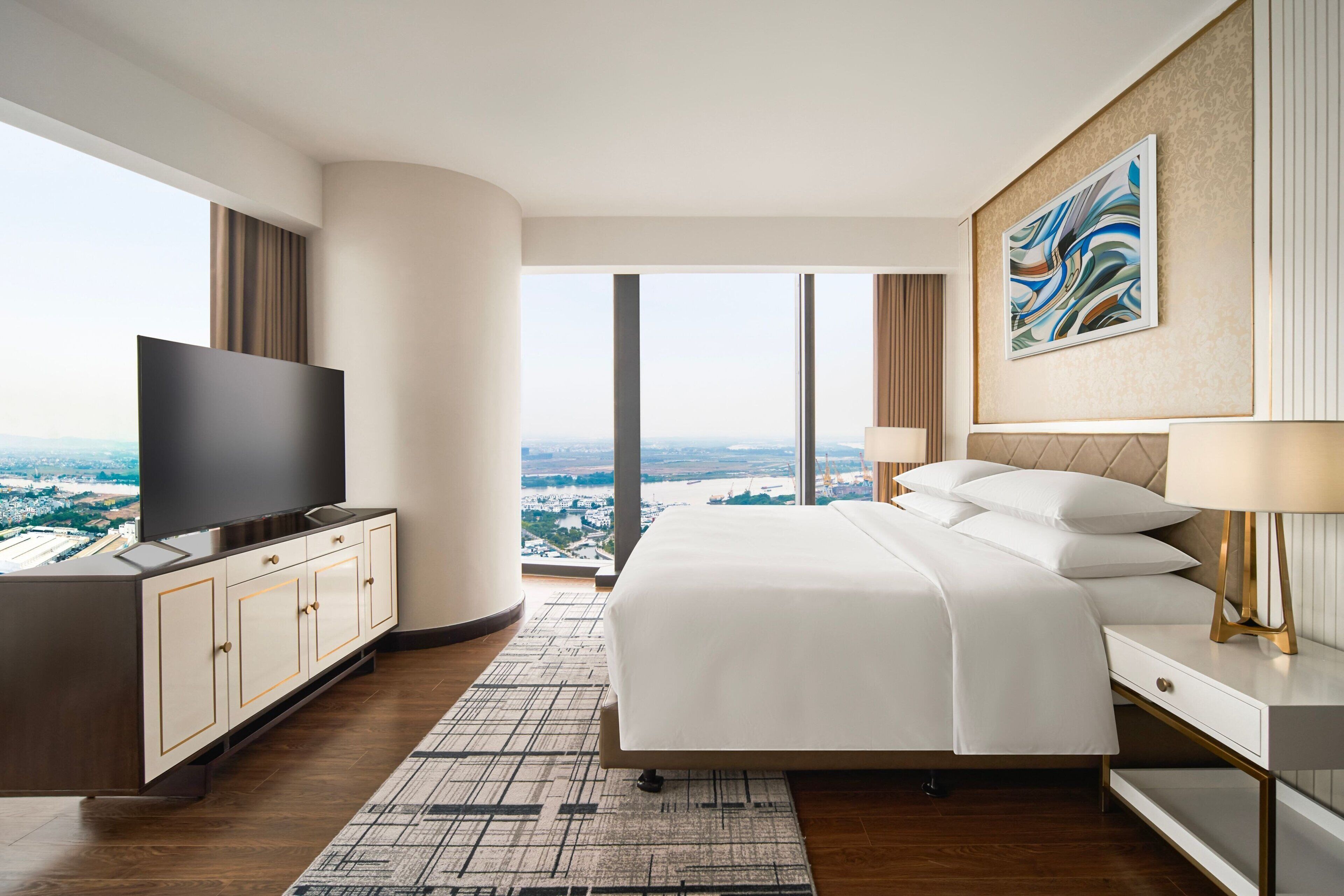 A modern hotel room with a large bed, a flat-screen TV, a desk, and a view of the city.
