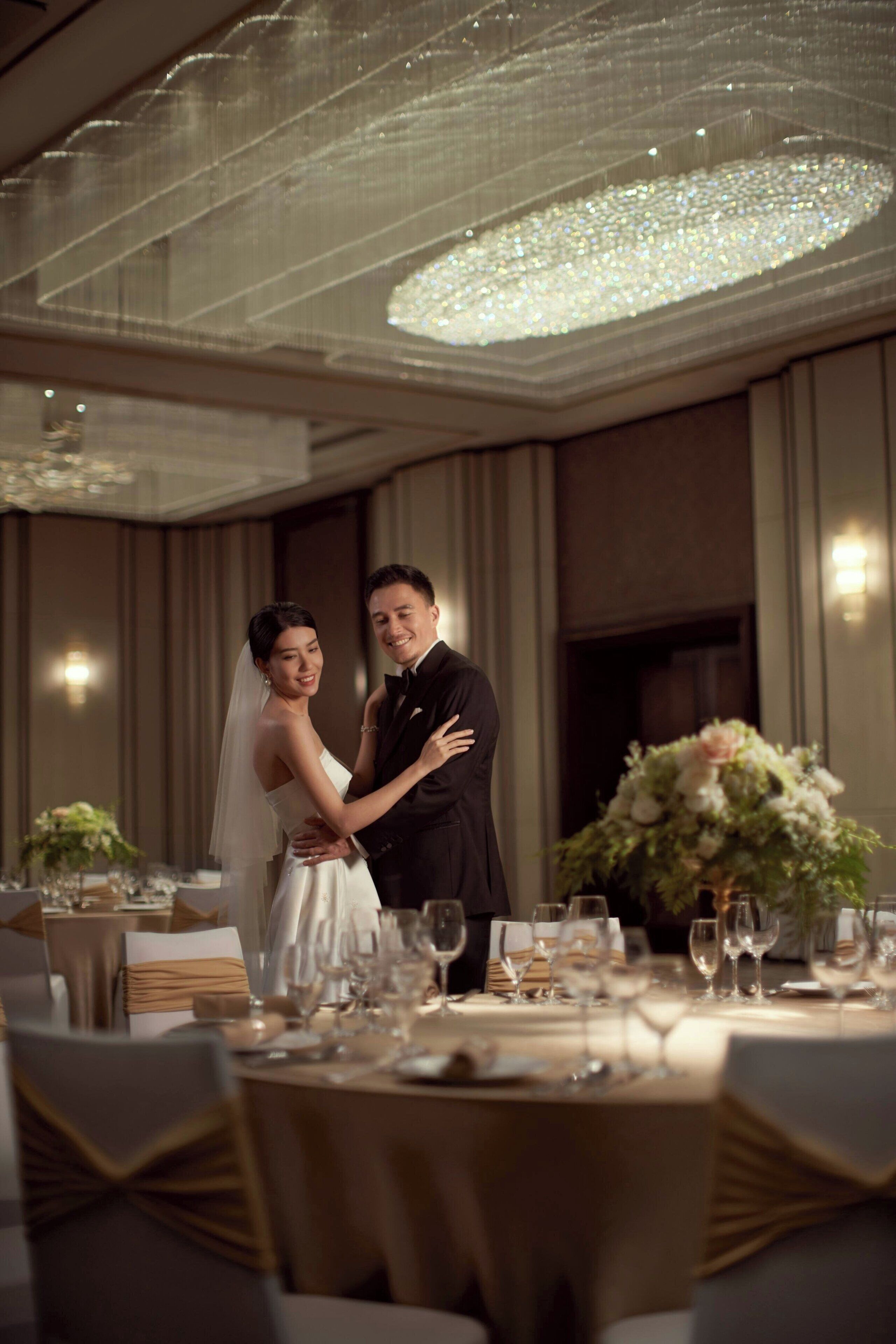A wedding reception with a couple embracing, a chandelier, and a table set for guests.