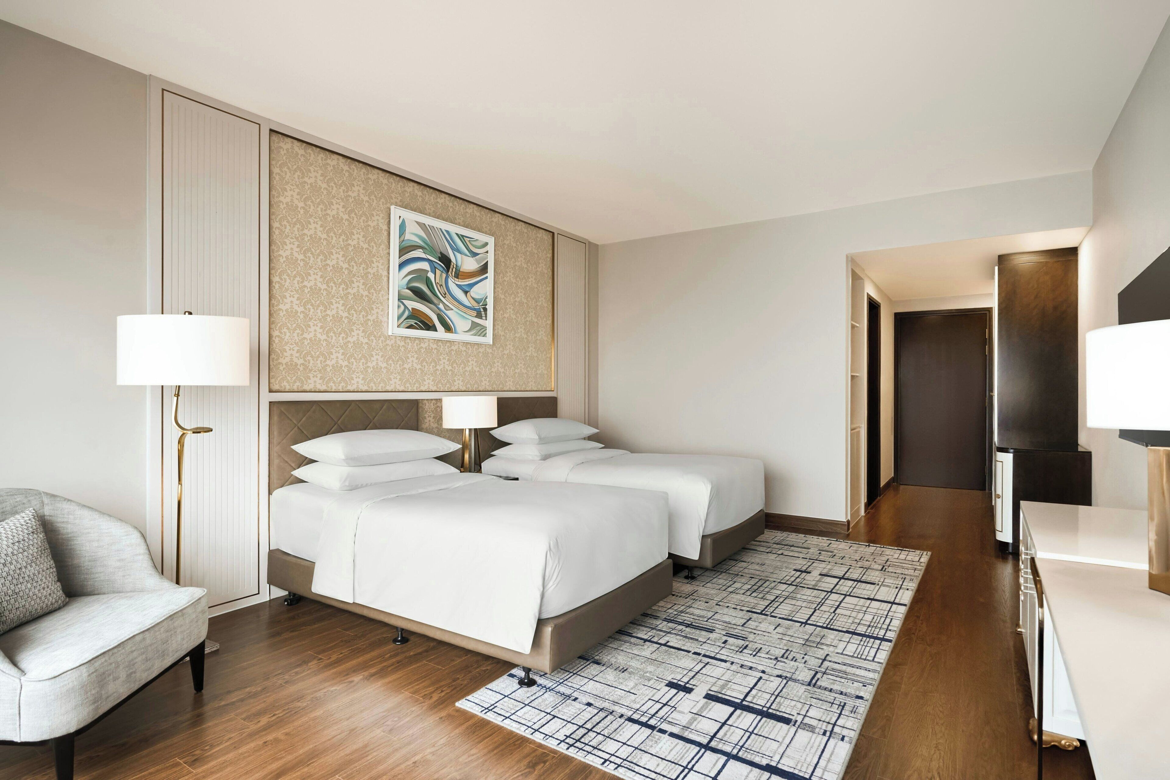 Una habitación de hotel moderna con una cama grande, dos mesitas de noche, un sofá, un televisor y una alfombra estampada.