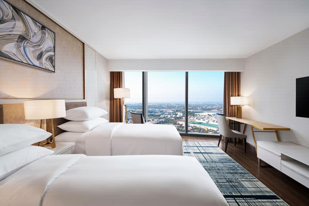 Ett modernt hotellrum med två sängar, ett skrivbord, en TV och ett stort fönster med utsikt över staden.