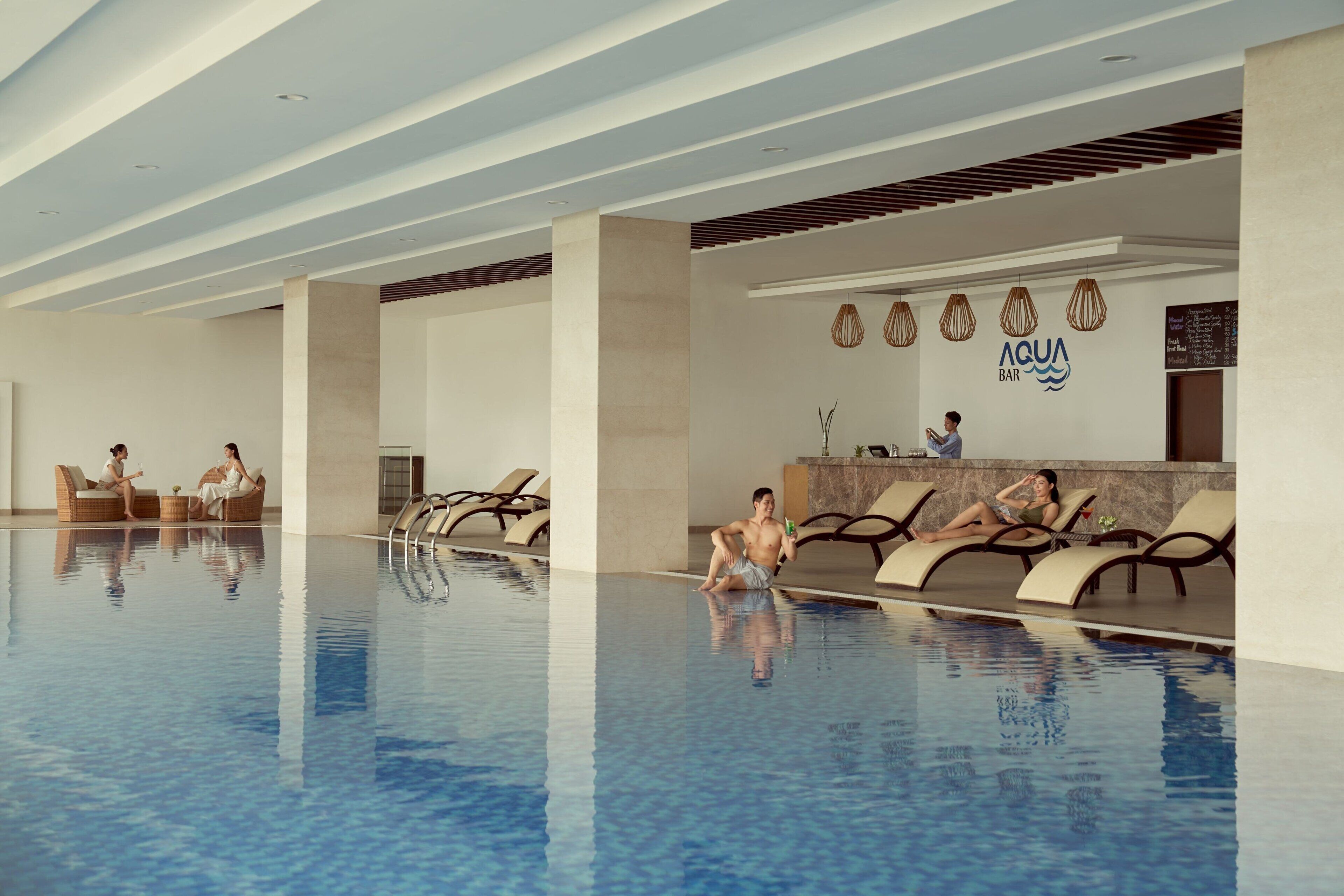 A modern indoor swimming pool with lounge chairs and a bar area.