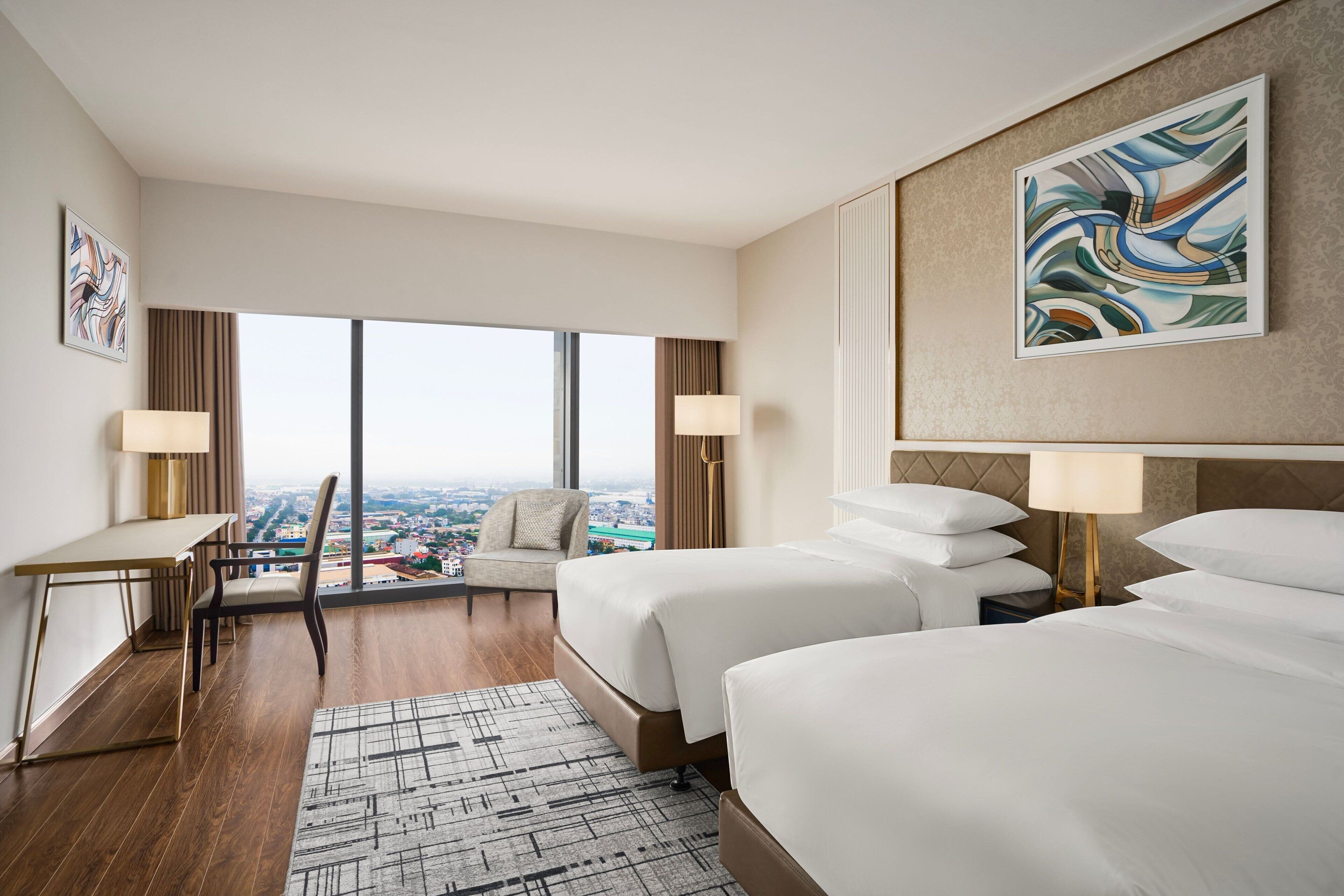 Een hotelkamer met twee bedden, een bureau, een stoel en een groot raam met uitzicht op de stad.