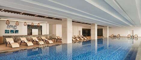 A modern indoor swimming pool with lounge chairs and a bar area.