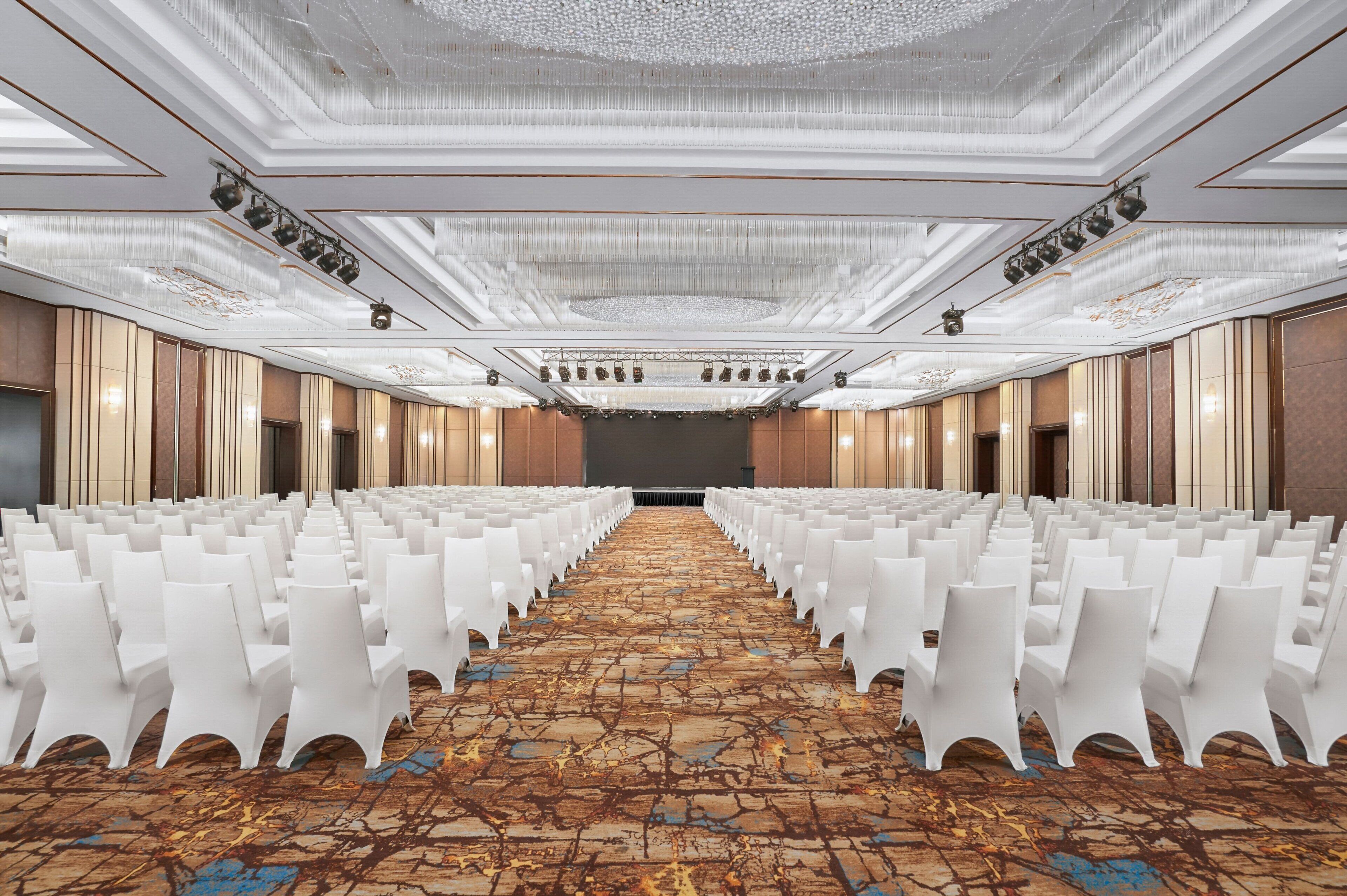 A conference room with rows of white chairs, a patterned carpet, and a high ceiling with chandeliers.