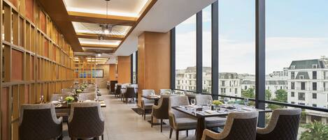 A modern dining area with high ceilings, large windows, and a view of buildings.