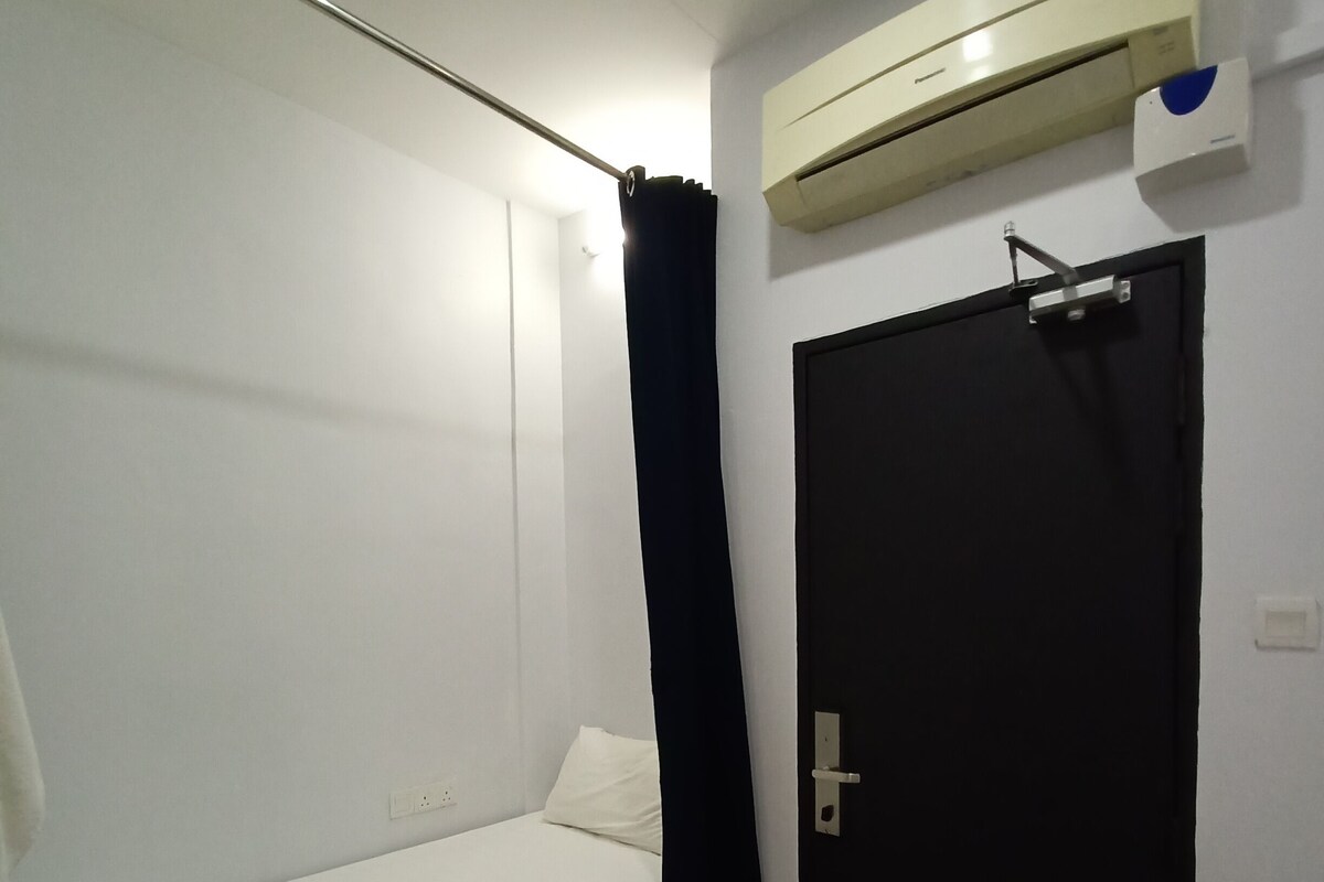 Une petite chambre avec un lit simple, une plinthe rouge, une porte noire et un appareil de climatisation fixé au mur.
