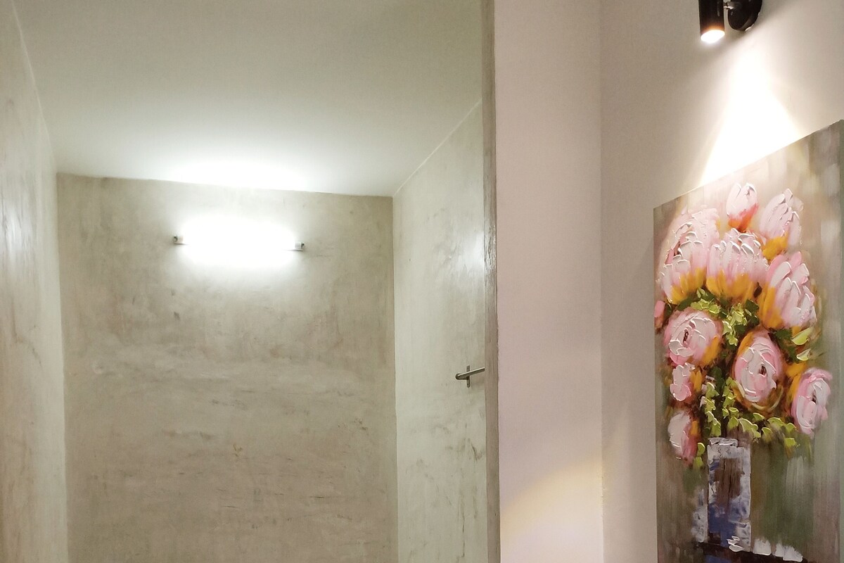 Une petite chambre avec un lit simple, un banc de rangement vert, un luminaire au plafond et un tableau floral accroché au mur.