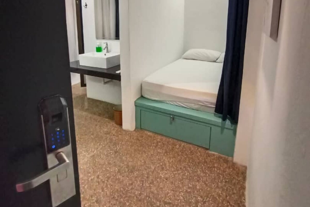 Une chambre d’hôtel de taille réduite, avec un lit simple, une petite salle de bain dotée d’un miroir et d’un lavabo, et une porte équipée d’une serrure numérique.