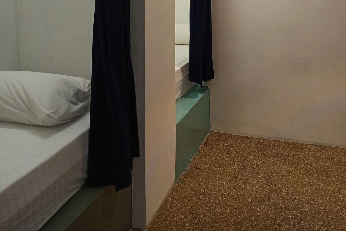 Une petite pièce faiblement éclairée, avec un lit simple, un rideau sombre et un appareil de climatisation fixé au mur.
