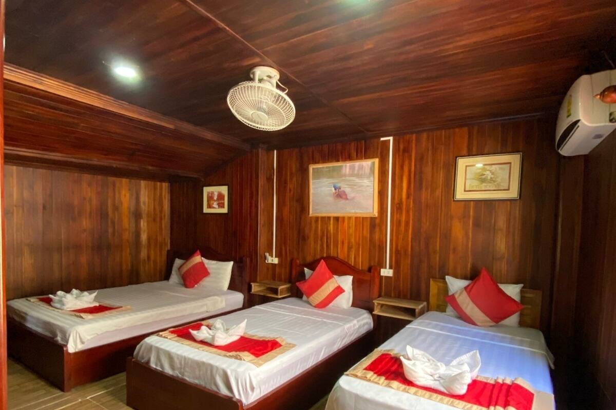 A room with four wooden beds, each with white bedding and red pillows. There is a ceiling fan, a wall-mounted air conditioner, and framed pictures on the walls.
