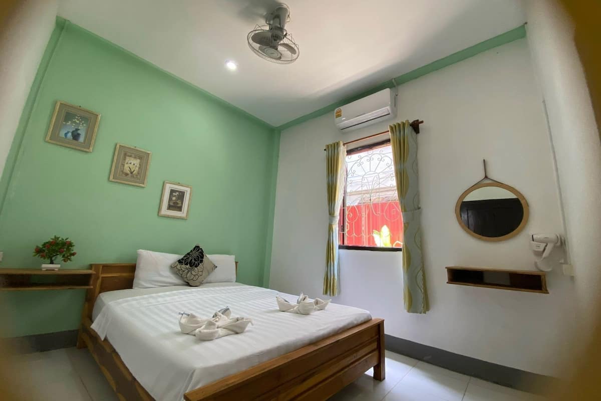 A bedroom with a bed, a bedside table with a plant, a window with curtains, a wall-mounted mirror, and an air conditioning unit.