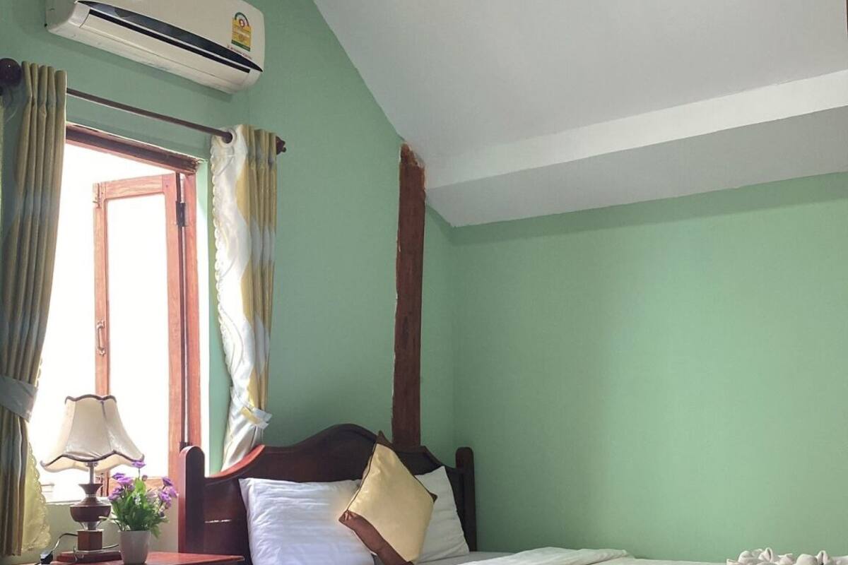 A hotel room with two beds, an air conditioning unit, a window with curtains, a bedside lamp, and a small table with a plant.