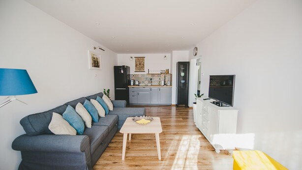 A modern living room with a grey sofa, wooden coffee table, and a yellow bench. A kitchen with grey cabinets and a TV is visible in the background. - Komplex Apartamenty Europa Sosnowiec (Sosnowiec)