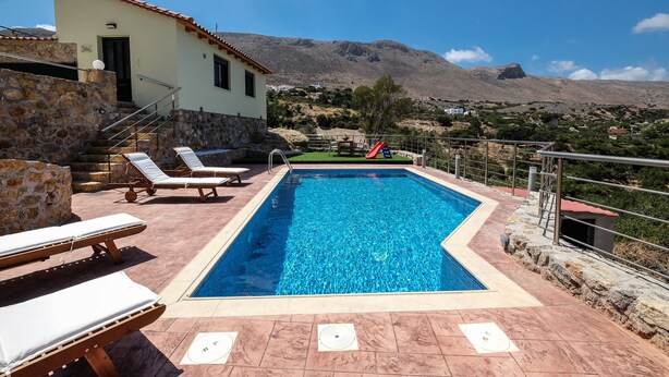 A swimming pool with lounge chairs, a stone building, and a mountainous backdrop. - Village chania (Platanias)