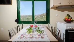 A dining table with a tablecloth, chairs, a window with a view of hills, a fruit bowl on a counter, and a television on the wall. - Village chania (Platanias)