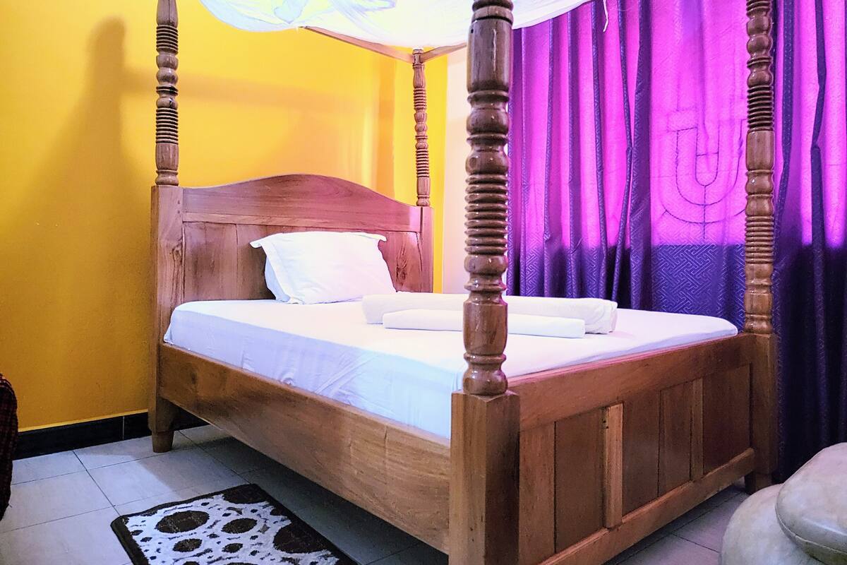 Un lit à baldaquin avec un couvre-lit blanc et des oreillers, une tête de lit en bois et un rideau violet en arrière-plan.