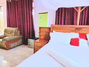A hotel room with a bed, a wooden nightstand, a chair, a small refrigerator, and a door with a green frame. - J M Peace Lodge (Dar es Salaam)
