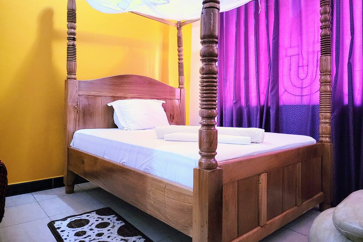 Un lit à baldaquin avec une structure en bois et une literie blanche, placé devant un mur jaune et agrémenté de rideaux violets.