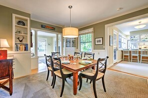 Un comedor con una mesa de madera, seis sillas y una araña. Se observan estanterías y una cocina al fondo.