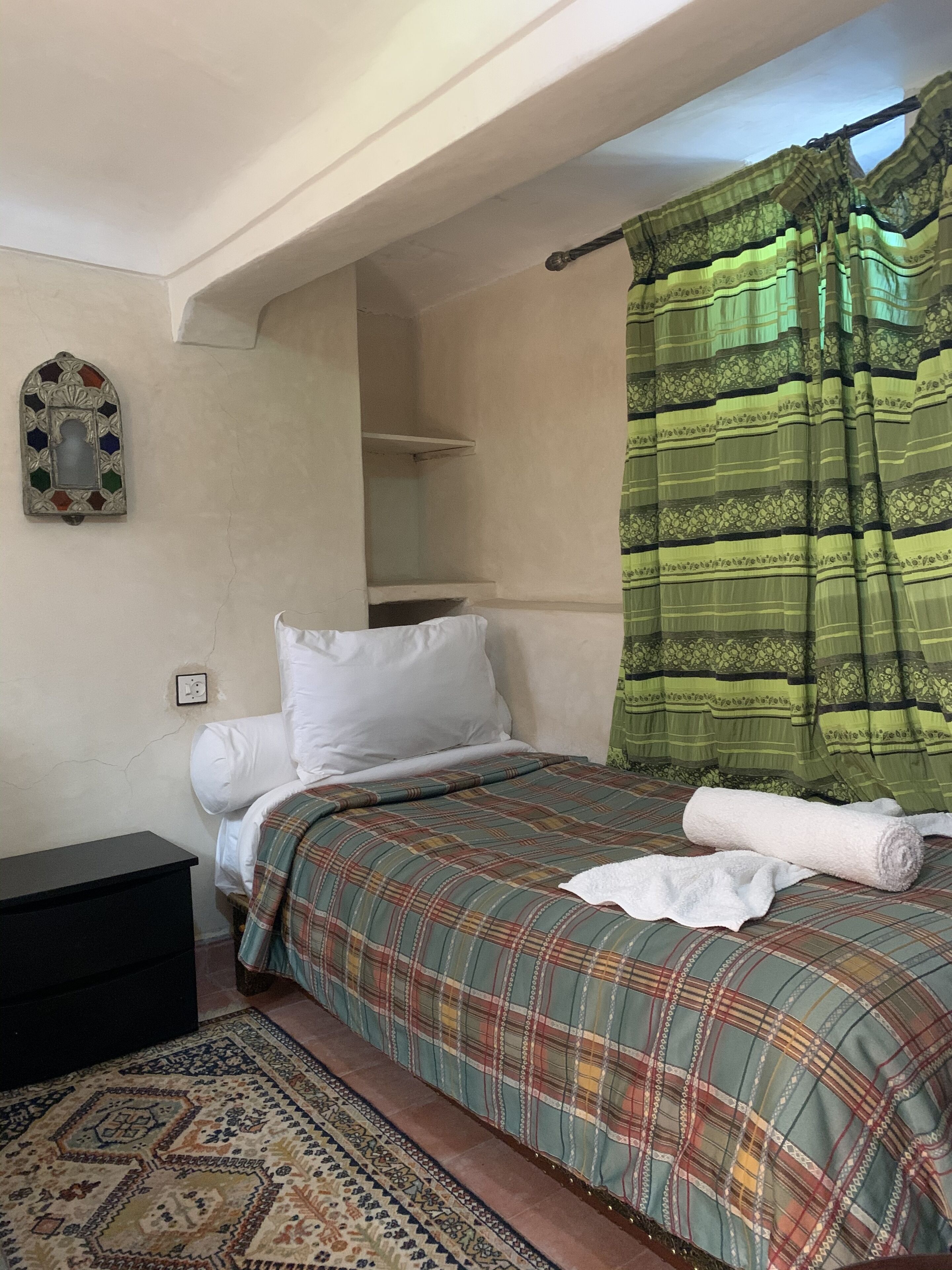 A bedroom with a bed, a nightstand, a patterned rug, a decorative mirror, and a green curtain.