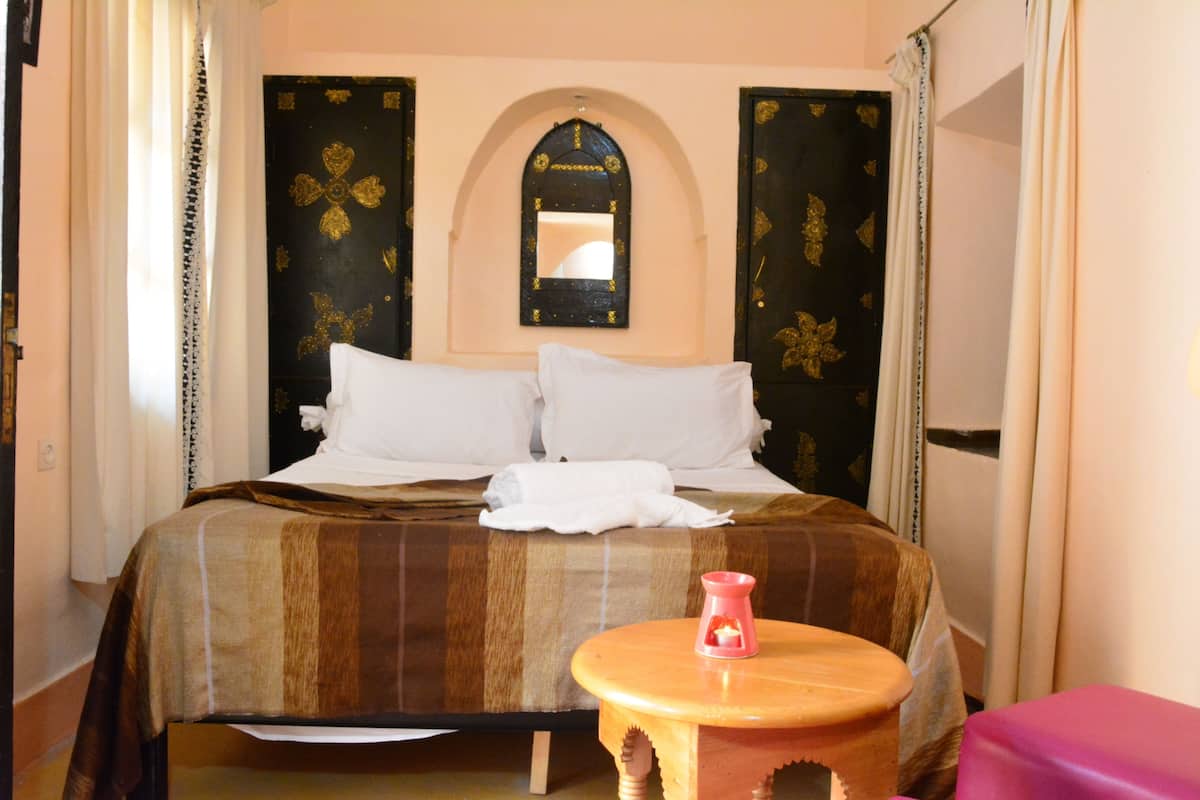 A hotel room with a bed, a small wooden stool, a mirror, and a door with a decorative pattern.