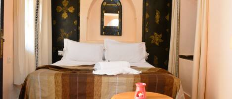 A hotel room with a bed, a small wooden stool, a mirror, and a door with a decorative pattern.