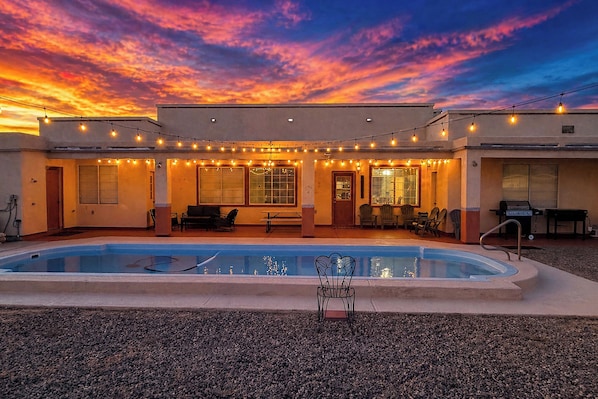 A pool area with string lights, a barbecue grill, and a patio with chairs and tables.