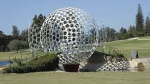 Escultura esférica com padrão de furos, cercada por área ajardinada com caminho e árvores.