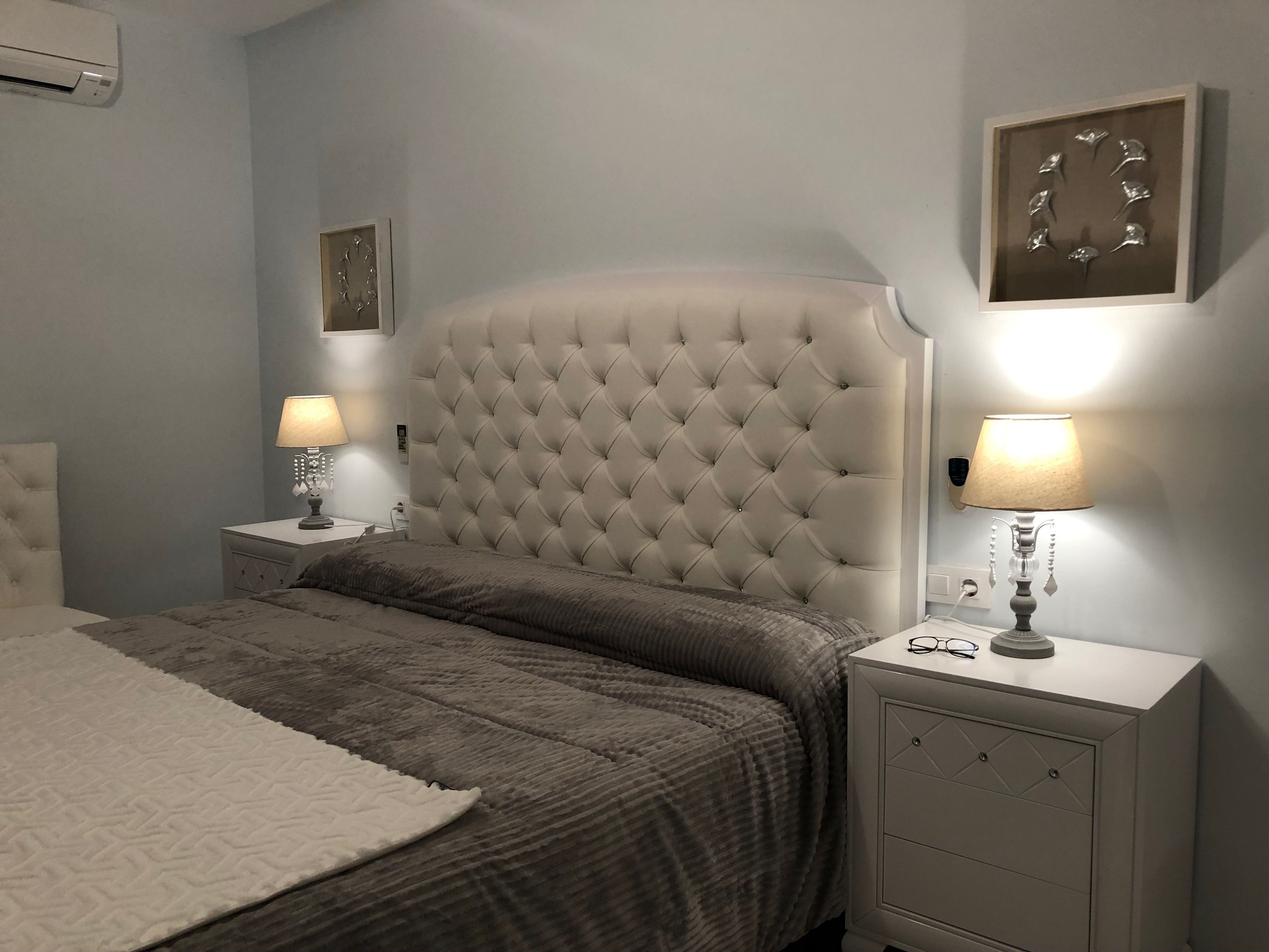 A bedroom with a tufted headboard, a white bedside table with a lamp, a bedside table with a lamp, a white rug, and a wall clock.