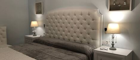 A bedroom with a tufted headboard, a white bedside table with a lamp, a bedside table with a lamp, a white rug, and a wall clock.