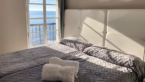 Uma cama com um edredom cinzento e duas toalhas enroladas, uma cabeceira branca e vista para o mar através de uma grande janela em arco.