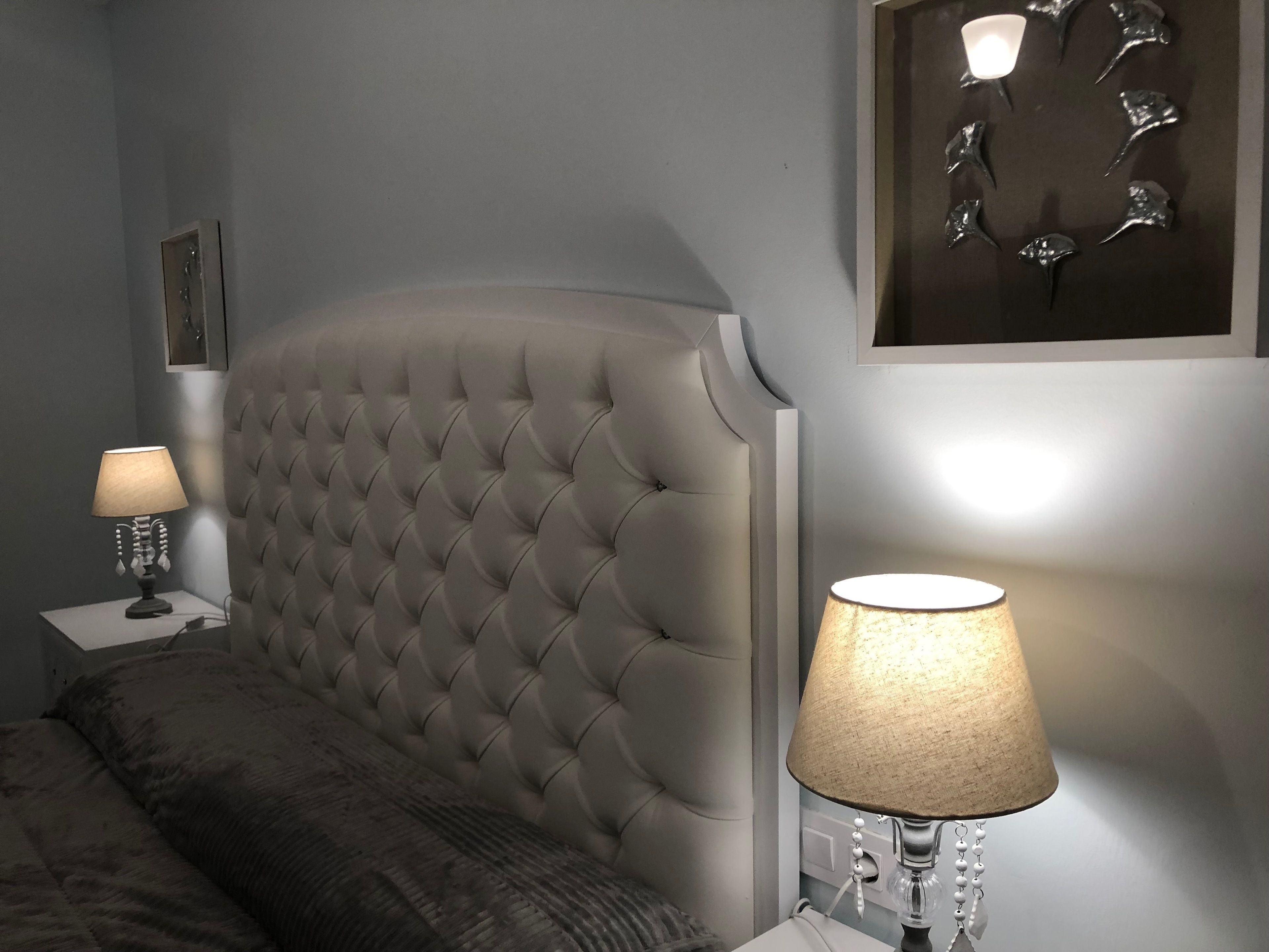 Un dormitorio con cabecera capitonada, mesita de noche con lámpara, y aplique de pared.