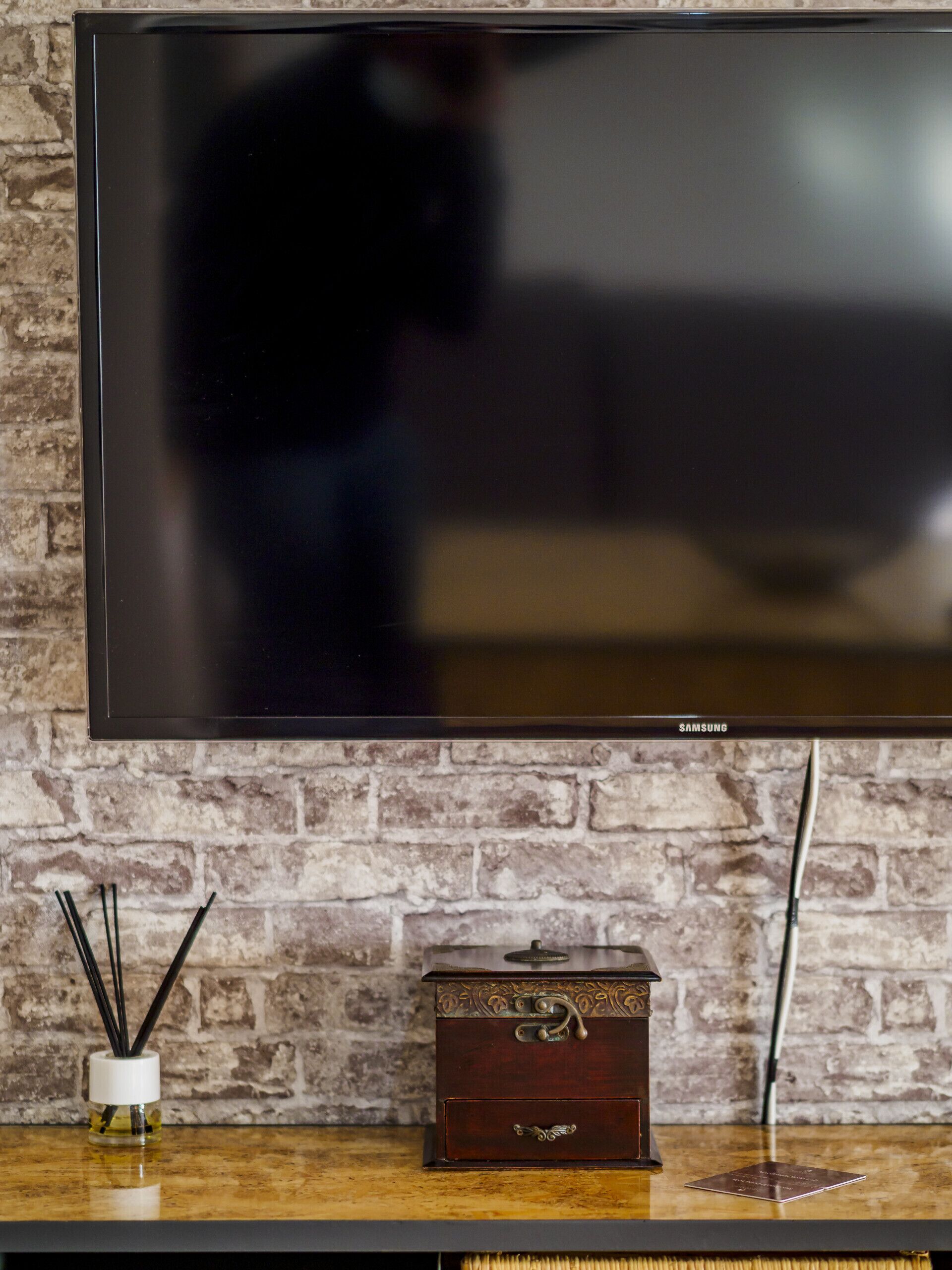 Un televisor de pantalla plana montado en una pared de ladrillo, un pequeño cofre de madera con un tirador decorativo, y un recipiente de vidrio con cañas de juncos sobre una superficie de madera.