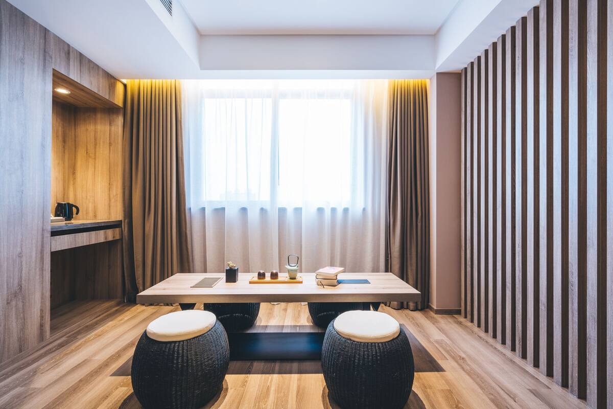 A modern room with a wooden floor, a long table with stools, a window with curtains, and a wooden panel wall.