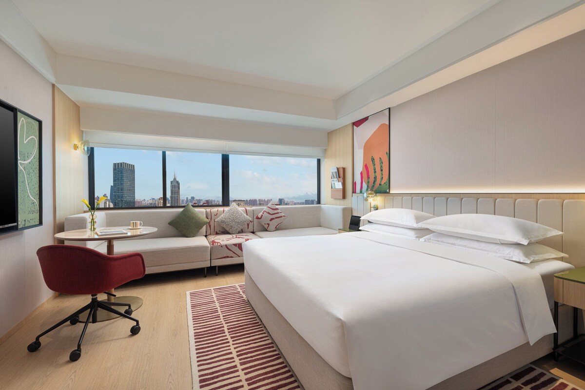 A modern hotel room with a large bed, a sofa, a desk, and a chair. There is a large window with a city view.