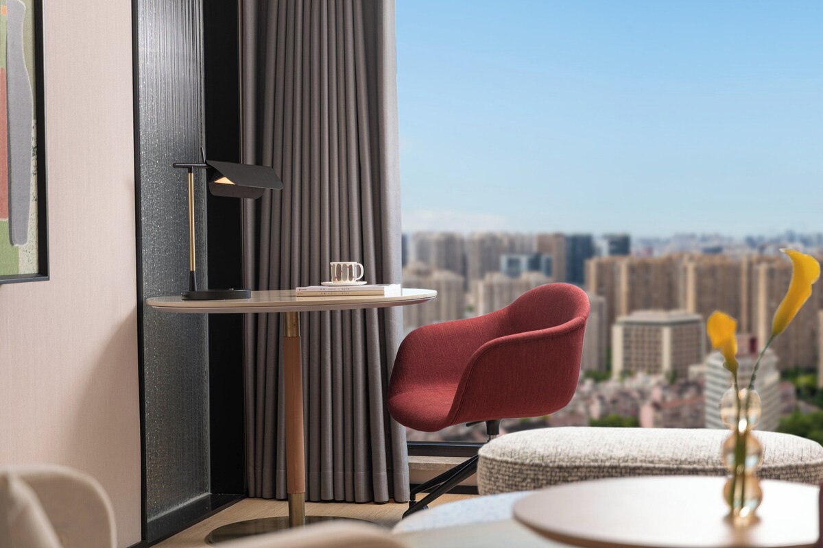 A modern room with a city view, featuring a red chair, a small table with a book and a vase of flowers.