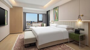 Una habitación de hotel moderna con una cama grande, una mesita de noche con lámpara, vistas a la ciudad y un televisor de pantalla plana de gran tamaño.