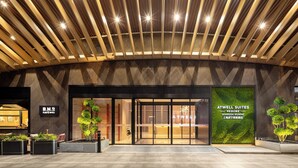 Entrada moderna de un hotel con techo de madera curvada, puertas de cristal y un letrero vertical verde para Atwell Suites.