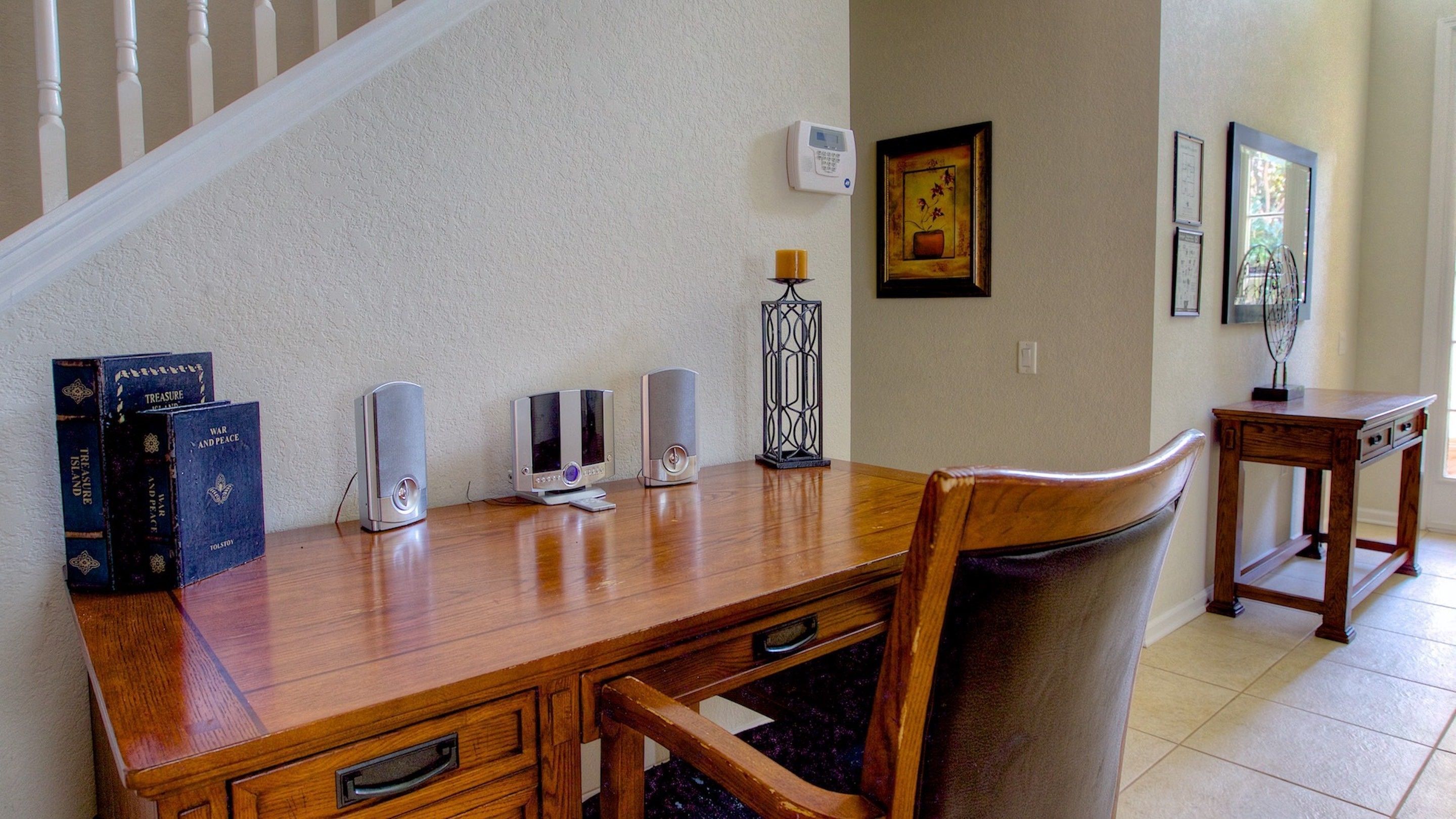 A wooden desk with books, a computer, and a speaker in a room with a staircase and framed pictures on the wall.
