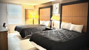 Habitación de hotel con dos camas, un escritorio, una silla, una lámpara y una ventana con persianas.