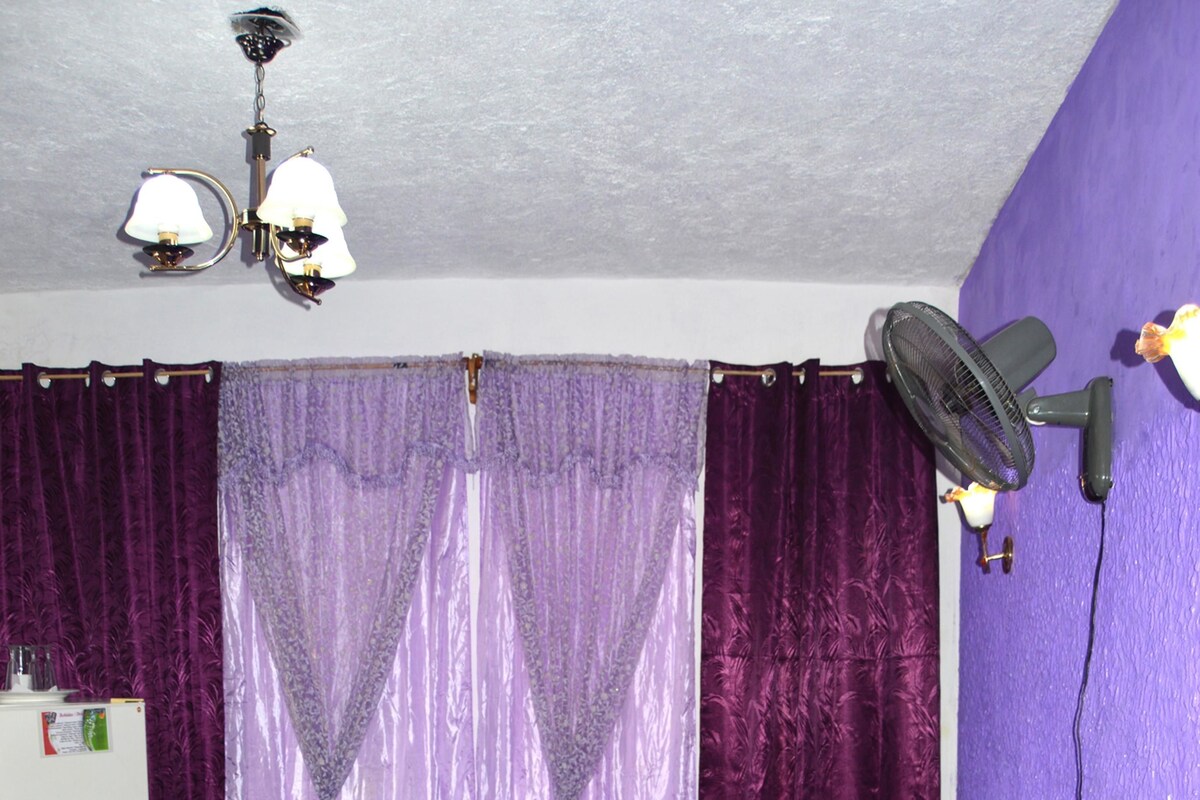 Un dormitorio con cama, cortinas moradas, ventilador de techo y refrigerador blanco.