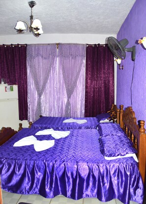 Un dormitorio con cama, cortinas moradas, ventilador de techo y refrigerador blanco.