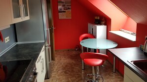 Eine kleine Küche mit roter Wand, ein Essbereich mit rundem Tisch und roten Stühlen sowie ein weißes Schranksystem mit einer grünen Tasche obenauf.