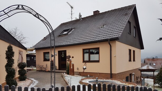 Ein zweistöckiges Haus mit Satteldach, einer Veranda und einem umzäunten Garten.