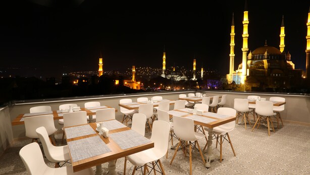 Zona de comedor en la azotea con mesas y sillas, con vistas a un paisaje urbano de edificios iluminados y una mezquita con minaretes.