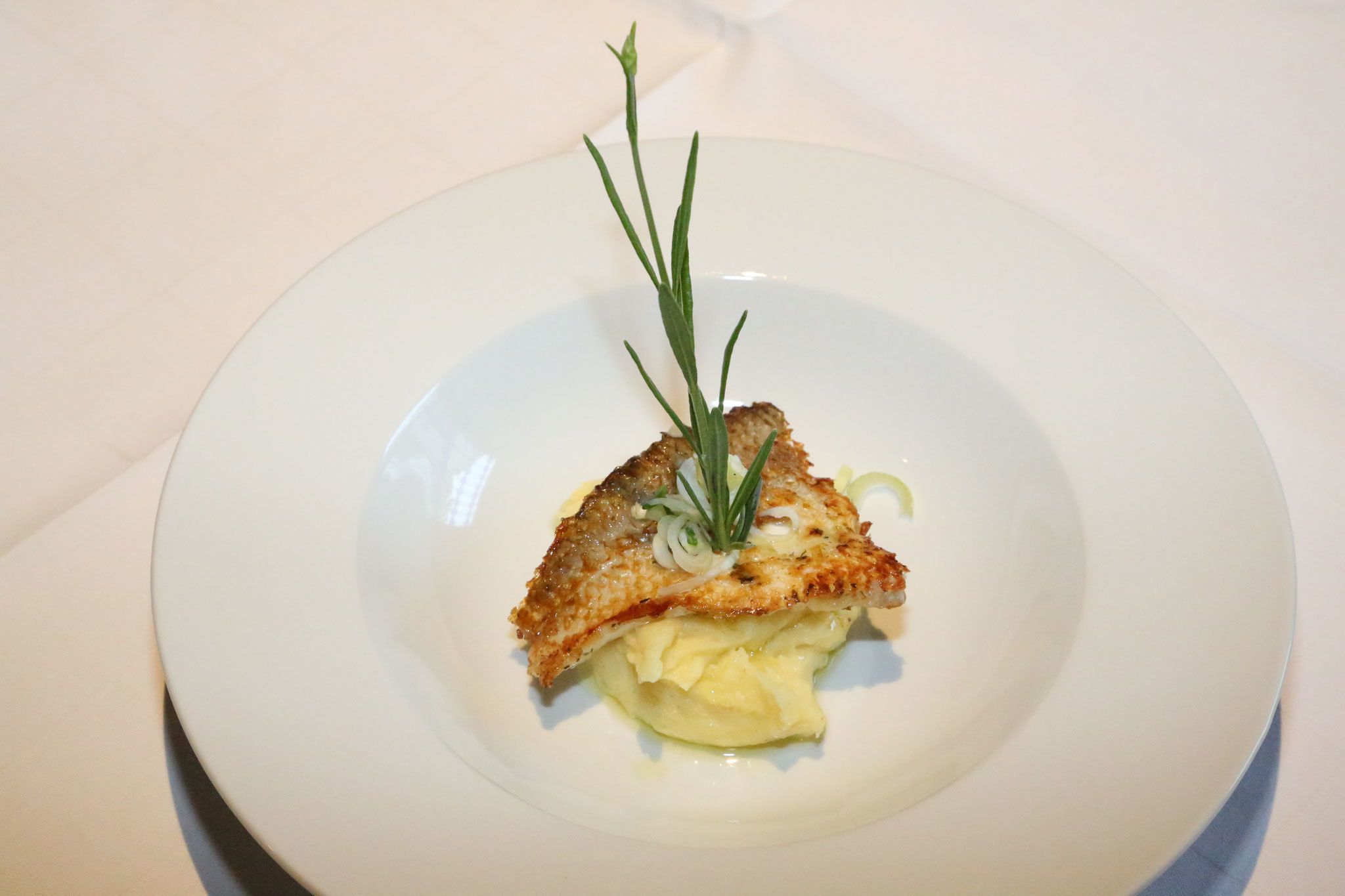 A white plate with a piece of breaded fish, garnished with a sprig of rosemary and a slice of lemon.