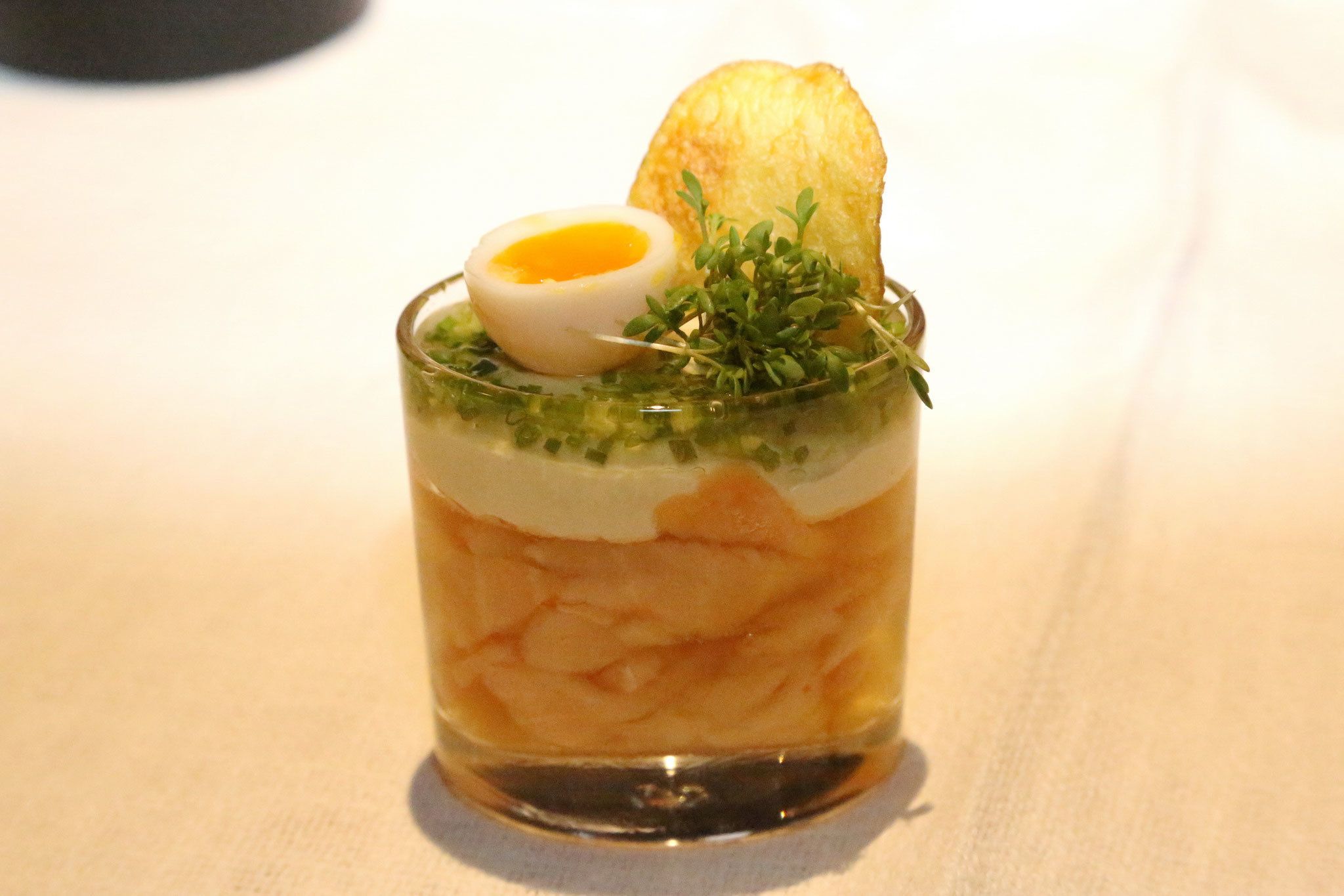 A glass containing a layered dish with a boiled egg, green garnish, and a piece of toast.