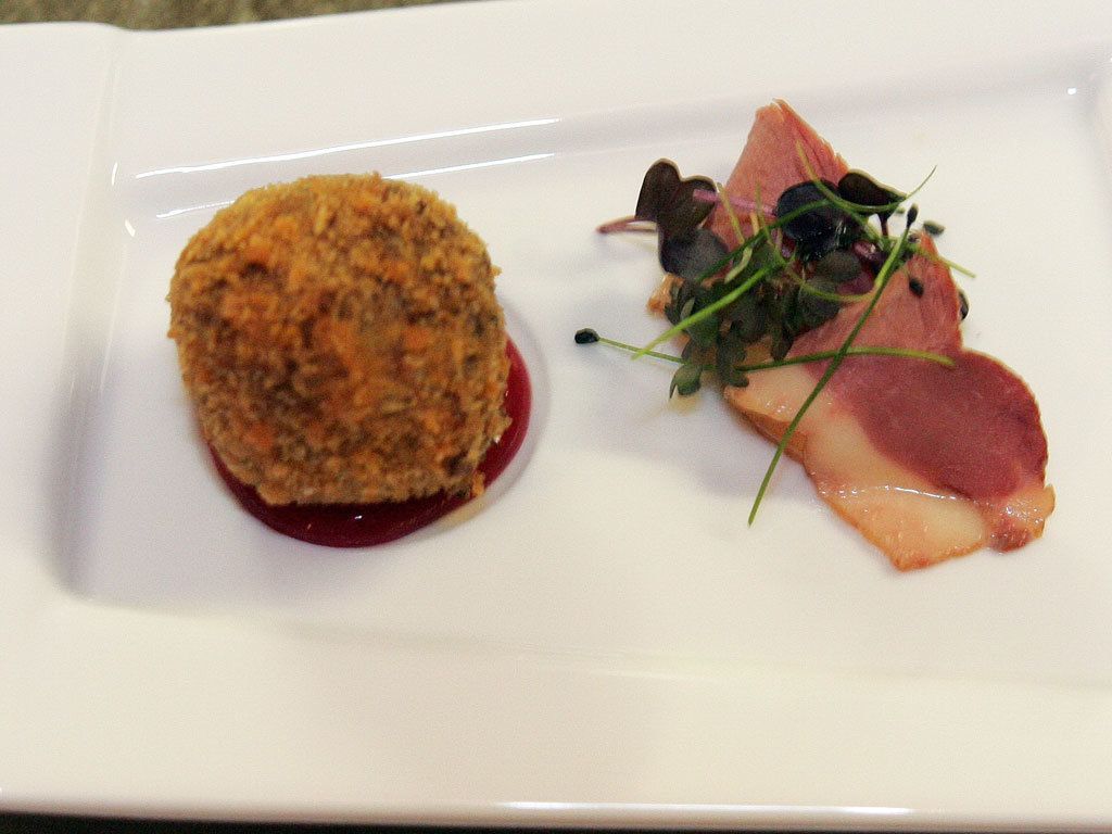 Ein weißer Teller mit einem runden, panierten Gericht und einer Fleischscheibe mit Salat.