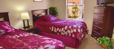 A bedroom with two beds, a nightstand with a lamp, a dresser, and a window with a view of palm trees.